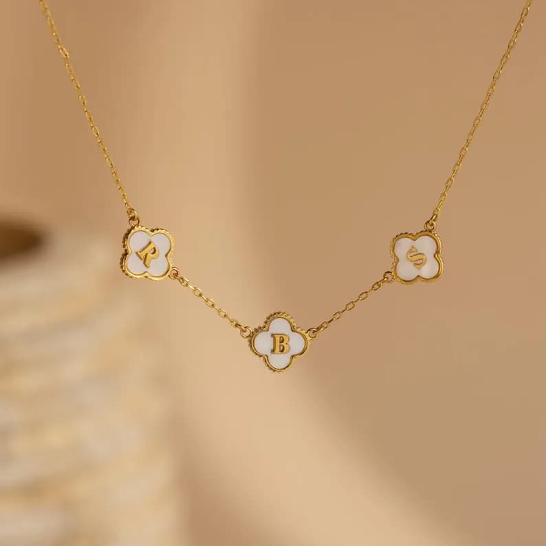 A gold chain necklace with three white clover charms, each with a gold letter, 'R', 'B', and 'S'.