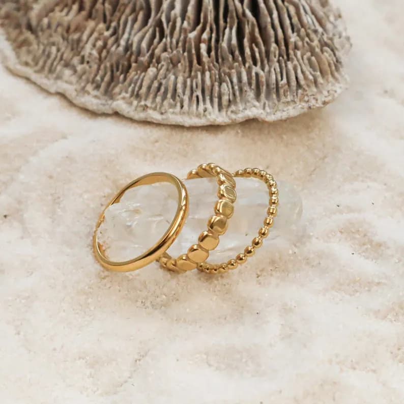 Three gold rings stacked on top of each other. The top ring is a beaded ring, the middle ring is a ring with square-shaped links, and the bottom ring is a simple band.