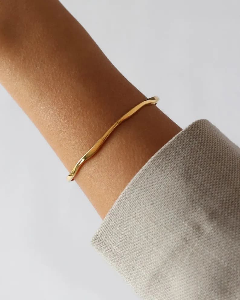 Gold-colored, wavy bangle bracelet worn on a person's arm, with a neutral-colored jacket in the background. Simple, elegant design.