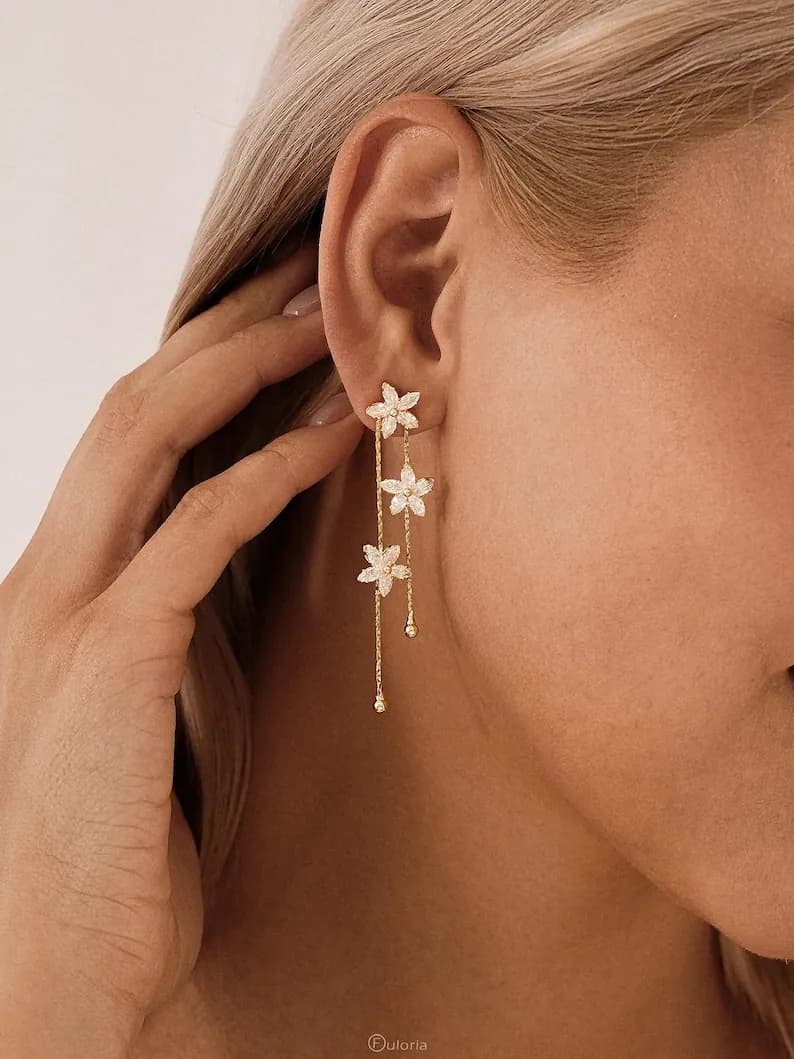 Gold-toned dangle earrings featuring three delicate, white flower pendants. The earrings have a linear design with small, dangling accents. The floral design adds a touch of elegance to the jewelry.