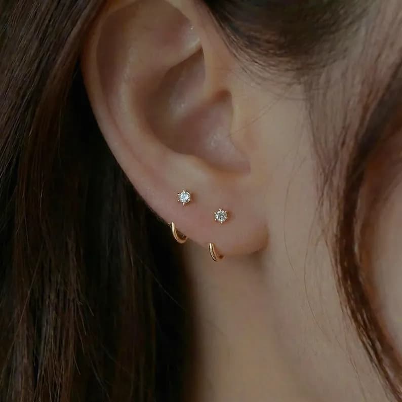 Close-up of an ear with two sets of earrings. One set features small, gold hoop earrings. The other set includes small, gold stud earrings with a single, clear gemstone.